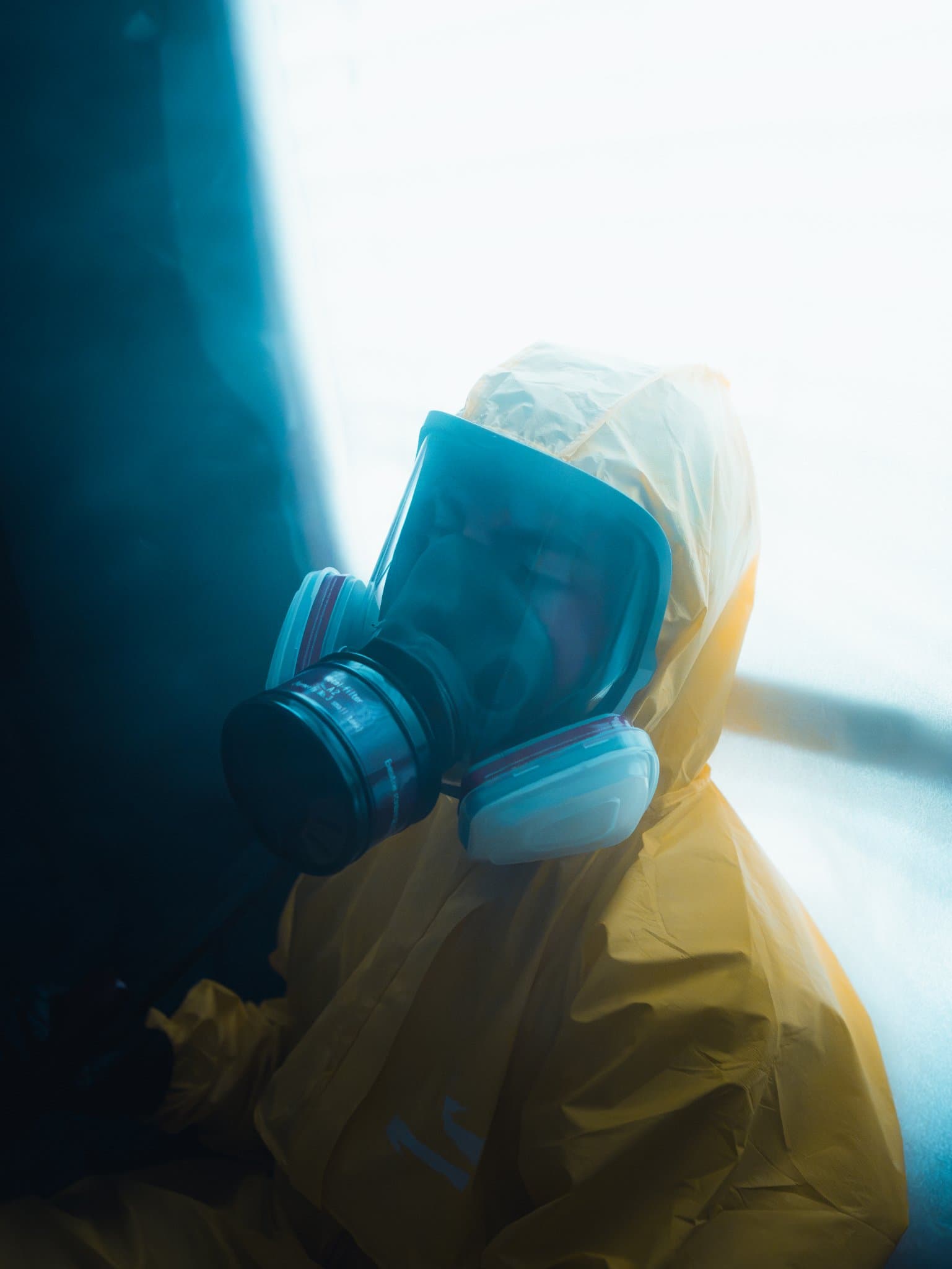 Tight profile close-up of a gas mask strapped over a yellow hazmat suit hood, against a dark teal background — human-made reference photo on Slow Sync