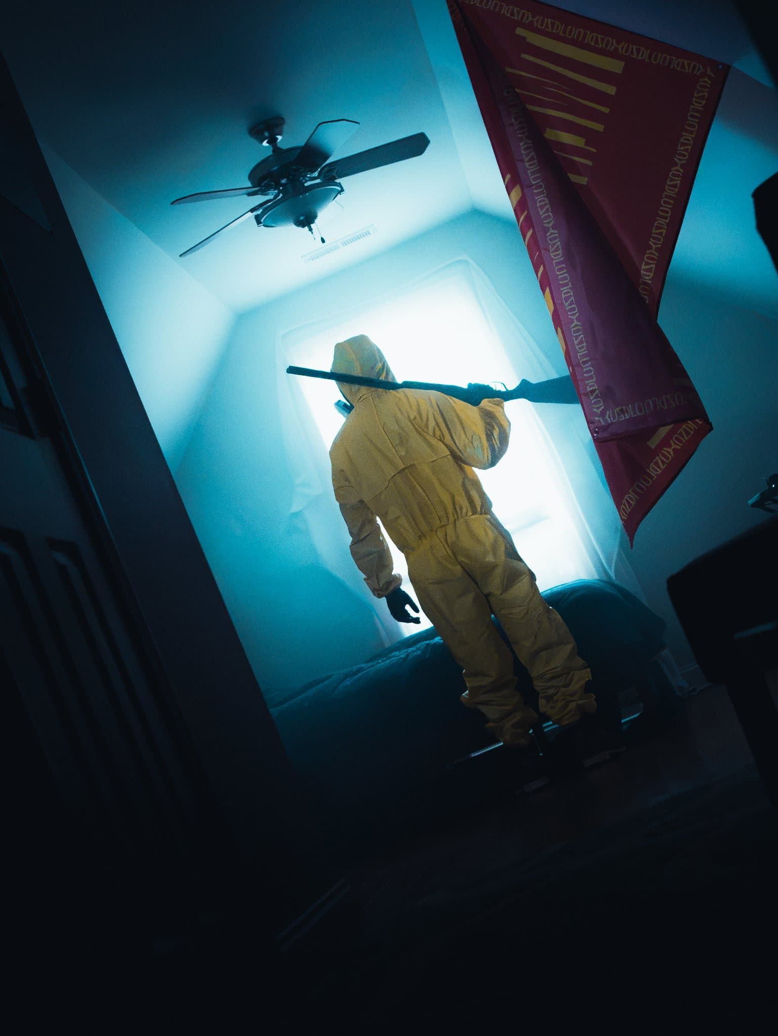 Figure in a yellow hazmat suit with a rifle over one shoulder, seen from behind in a teal-lit room with a red flag and a ceiling fan — human-made reference photo on Slow Sync
