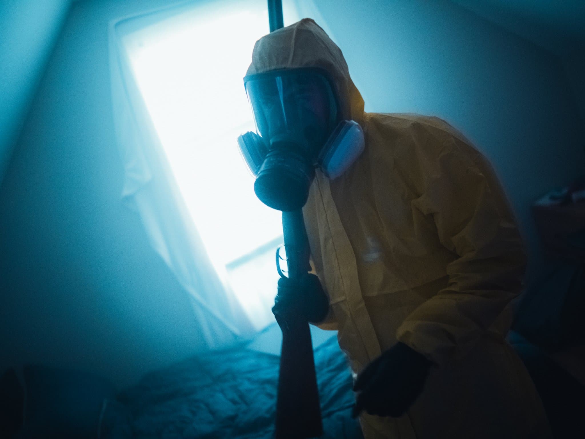 Person in a yellow hazmat suit and gas mask leaning toward the camera, holding a rifle upright, backlit by a bright window — human-made reference photo on Slow Sync