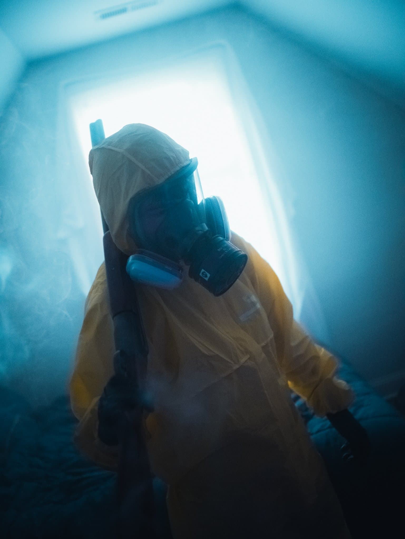Person in a yellow hazmat suit and gas mask holding a rifle down at their side, leaning through thin smoke — human-made reference photo on Slow Sync